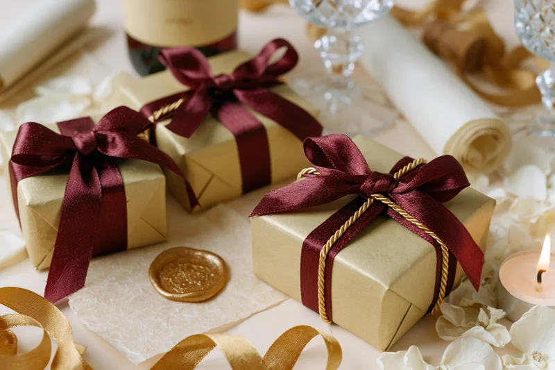 Gold gift boxes tied with burgundy ribbon and twisted gold twine on a decorated table with wax seal, candle, and flower petals