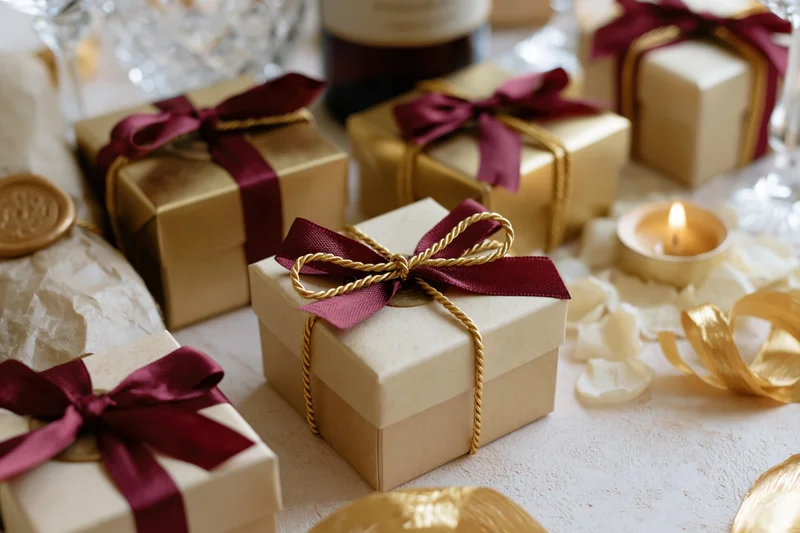 Gold gift boxes with burgundy ribbons and twisted gold string arranged on a table with candles and rose petals