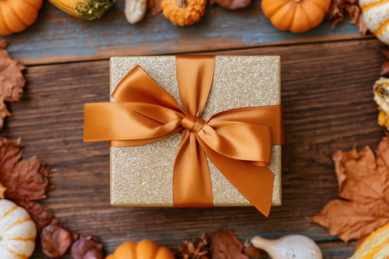 Gold glitter gift box with orange satin bow on wooden table surrounded by pumpkins and autumn leaves