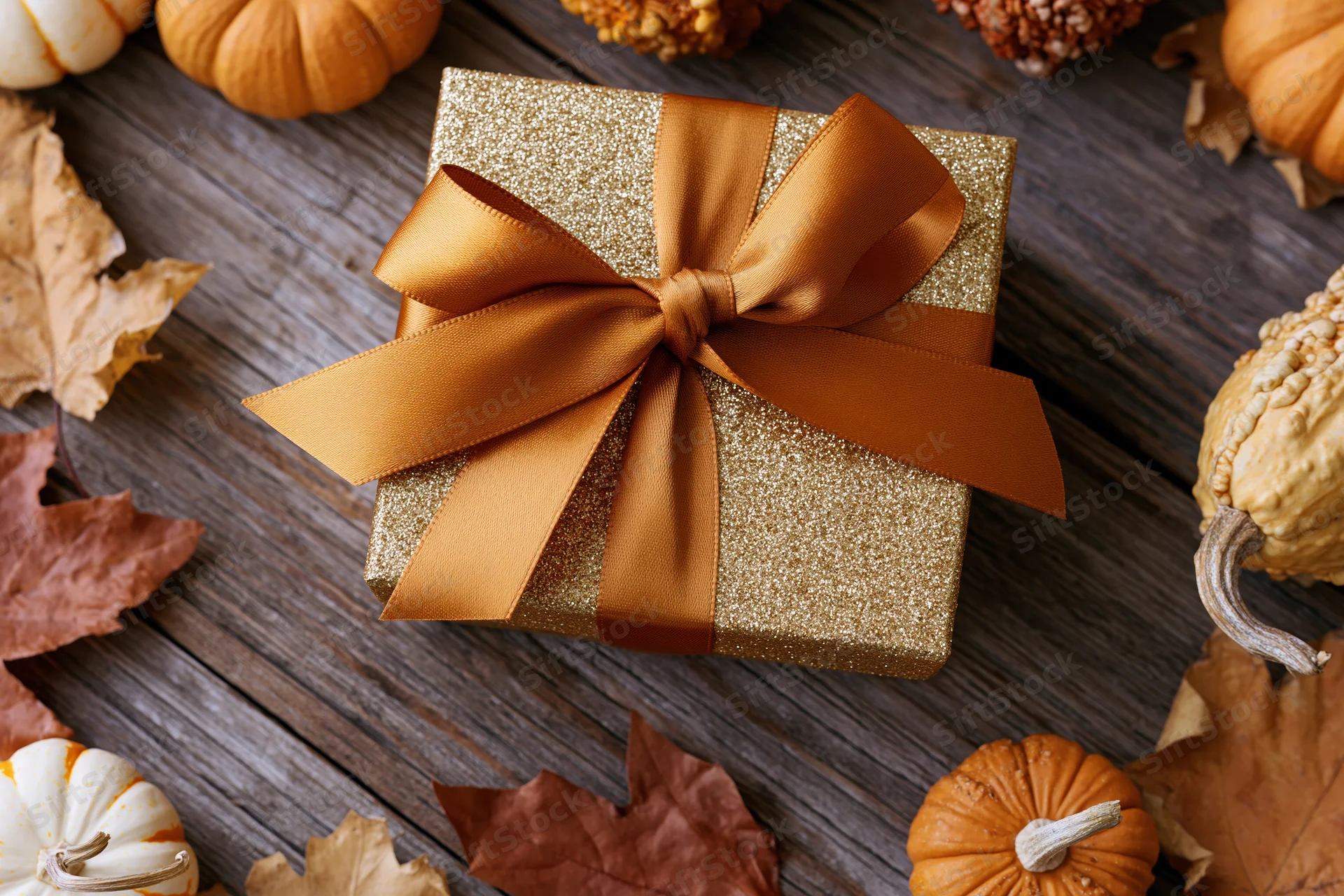 Gold glitter gift box with orange satin bow on wooden table surrounded by pumpkins and autumn leaves