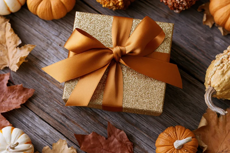 Gold glitter gift box with orange satin bow on wooden table surrounded by pumpkins and autumn leaves