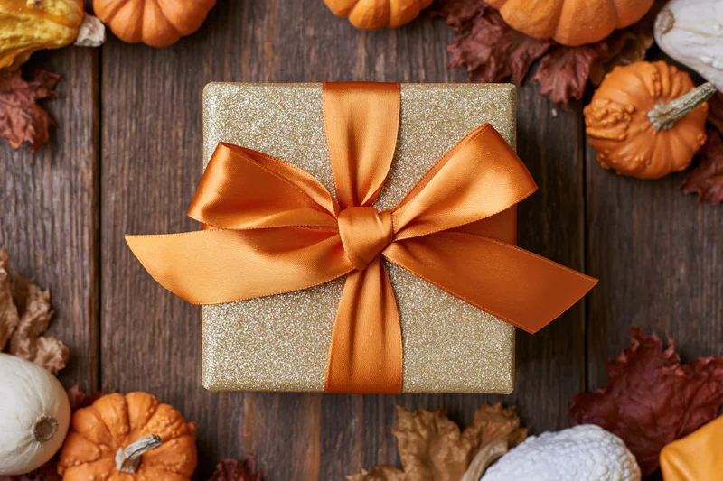 Gold glitter gift box with orange satin bow on wooden table surrounded by pumpkins and autumn leaves