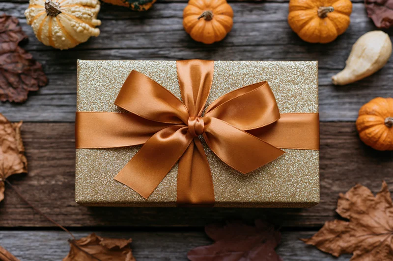 Gold glitter gift box with orange satin bow on wooden surface surrounded by pumpkins and autumn leaves