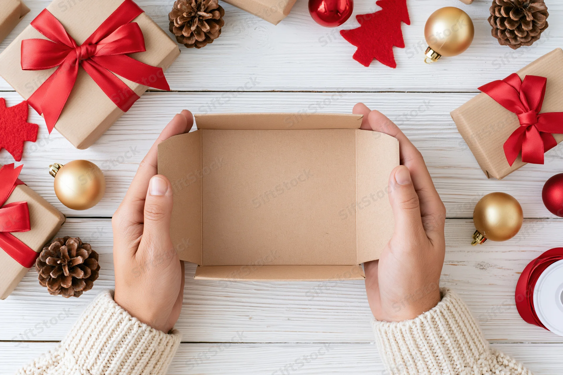 Hands holding open empty kraft gift box on white wooden table surrounded by christmas decorations