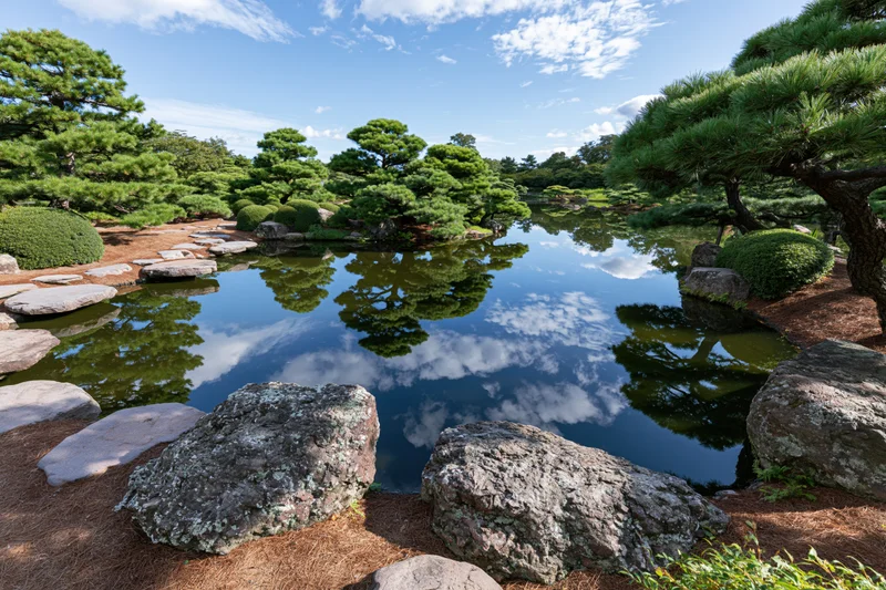 Japanese garden with calm pond reflecting sky and trees among rocks and pine trees