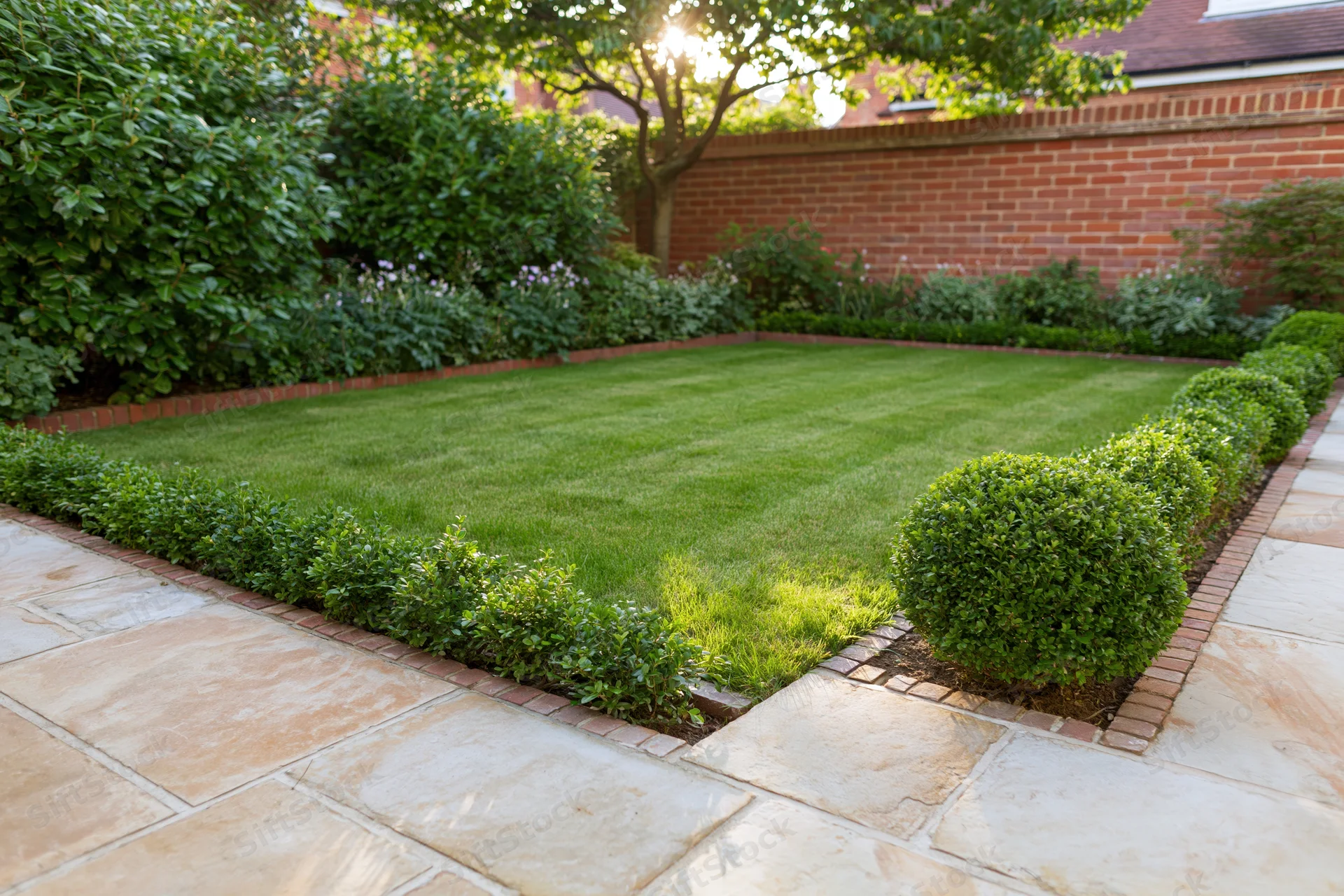 Manicured backyard garden with neatly trimmed grass lawn bordered by hedges and brick edging, surrounded by stone patio and brick wall