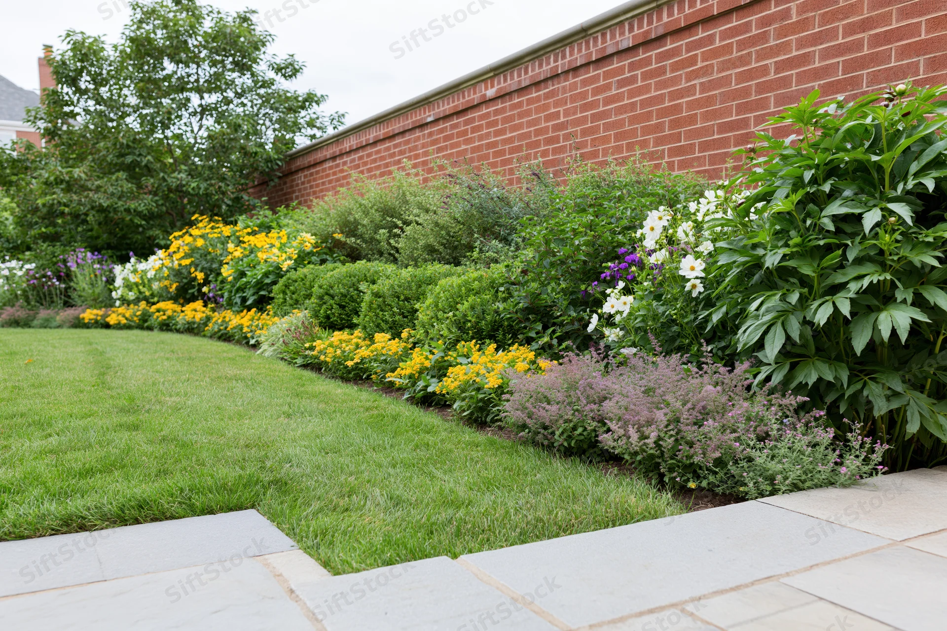 Manicured suburban backyard garden with neatly trimmed lawn and colorful flowerbeds along a brick wall and stone pathway