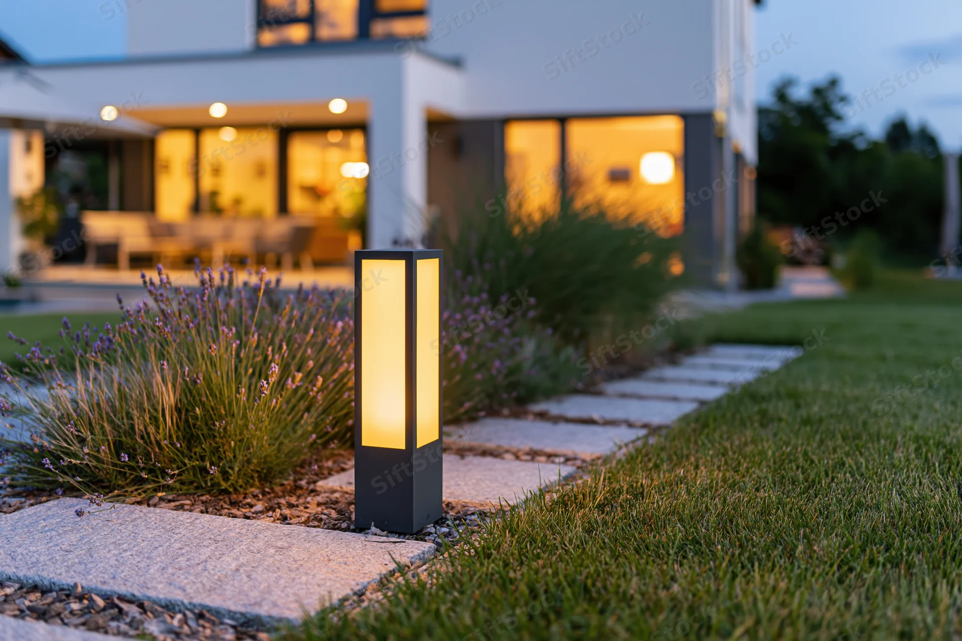 Modern suburban backyard at dusk with glowing bollard light along stone pathway beside lavender plants and house
