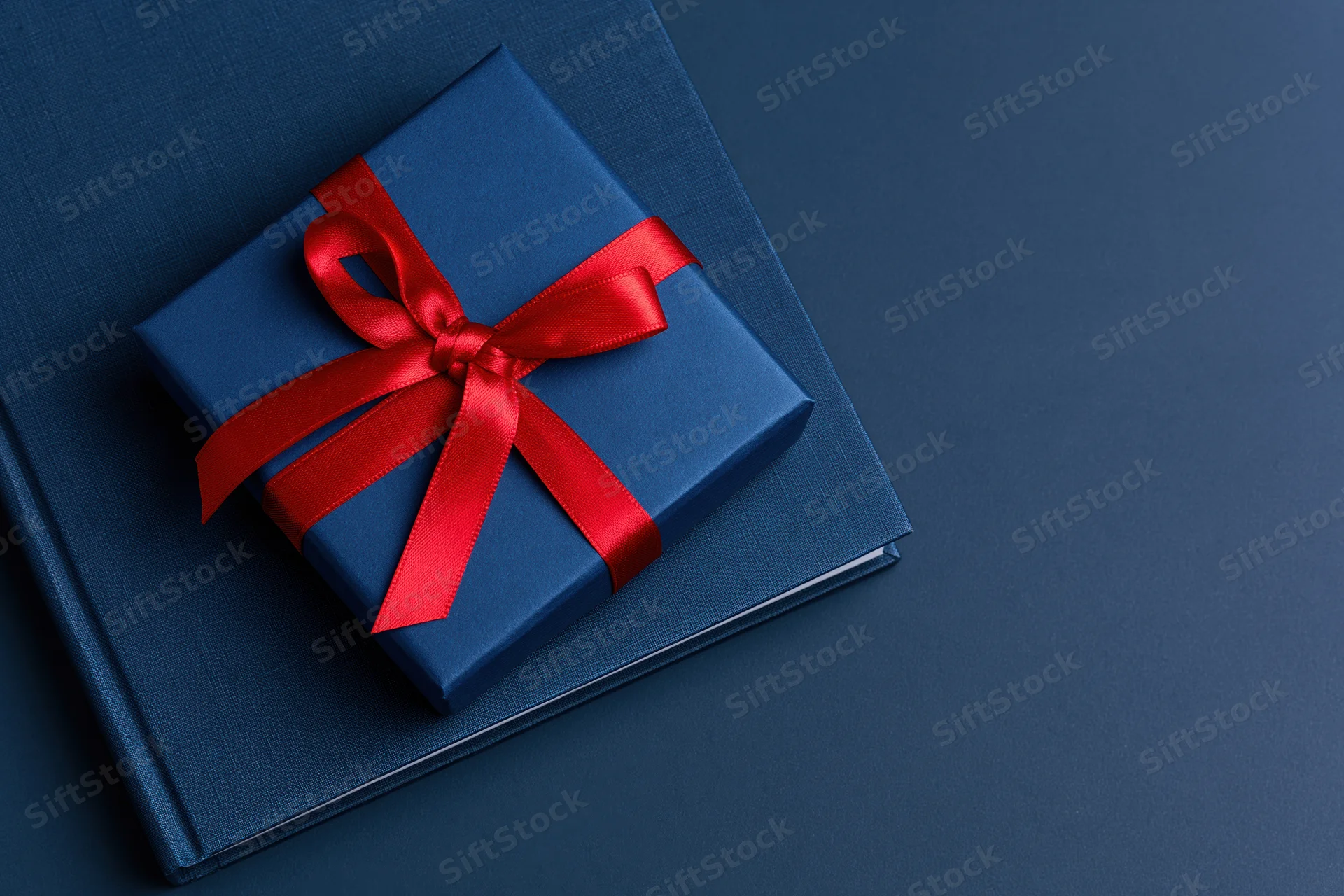 Navy blue gift box with red satin ribbon bow resting on dark blue textured surface