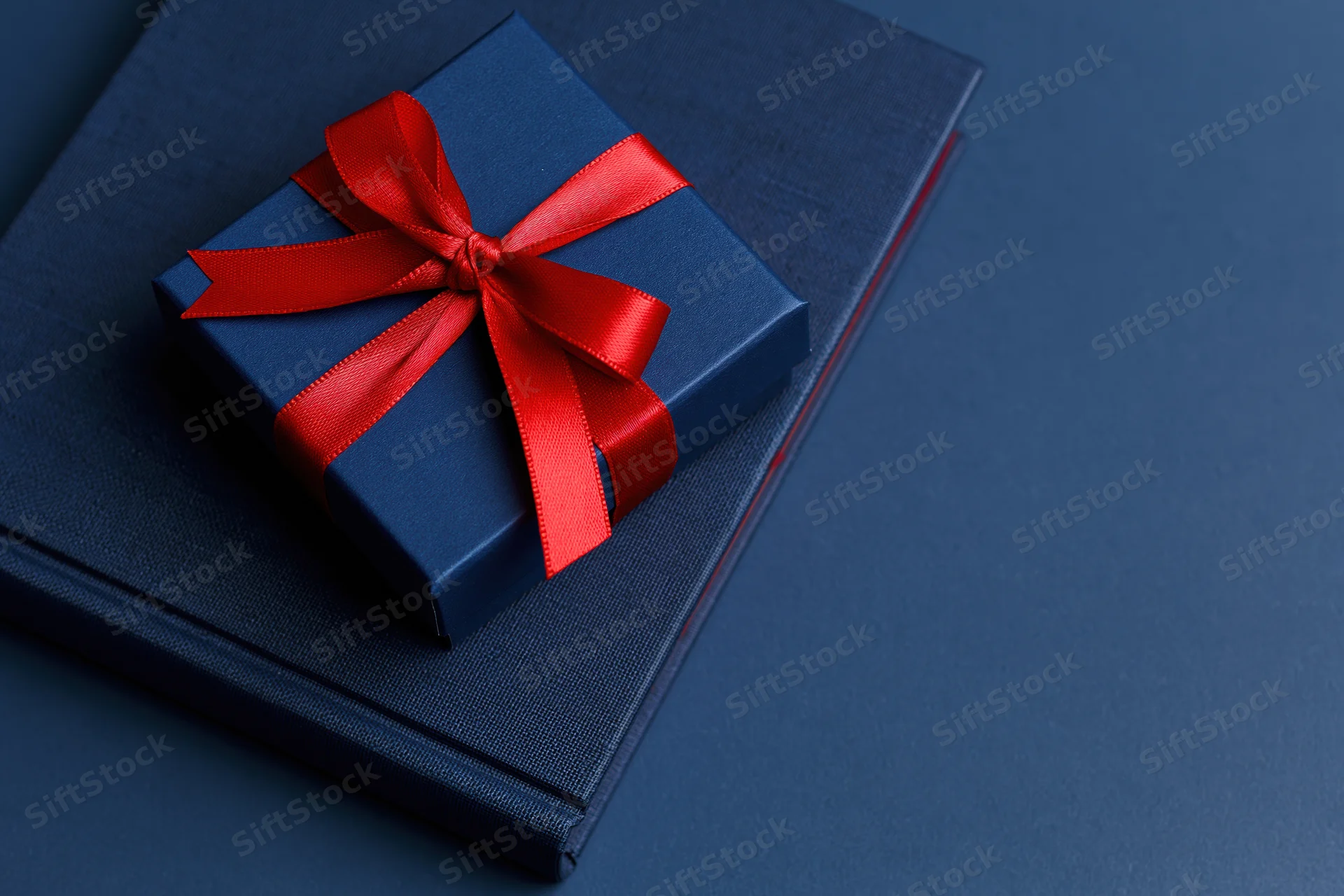 Navy blue gift box with red satin ribbon bow resting on a matching book on a dark blue surface