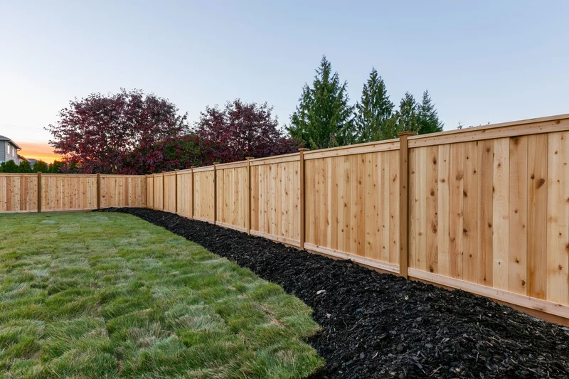 Wooden fence lining a newly sodded suburban backyard with mulch and trees at dusk