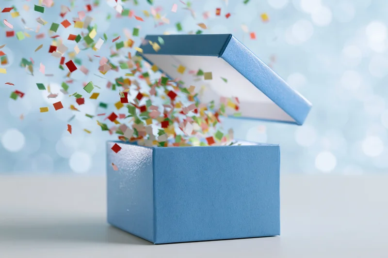 Open blue gift box with lid flying upward and multicolored paper confetti scattering out
