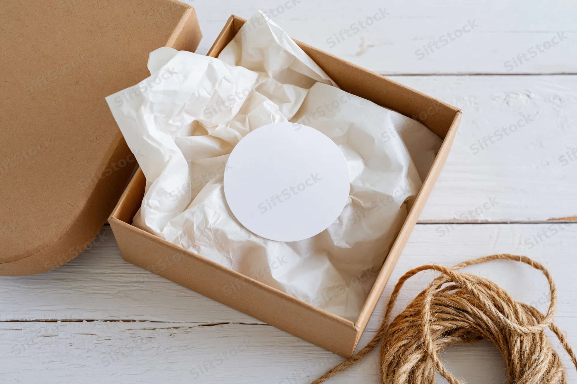 Open kraft cardboard gift box with crumpled paper and blank circular label inside, viewed from above on white wooden surface with twine beside it
