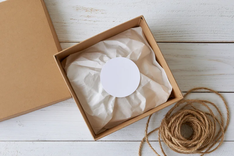 Open kraft cardboard box with white circular label on crumpled paper, twine beside, on white wooden surface