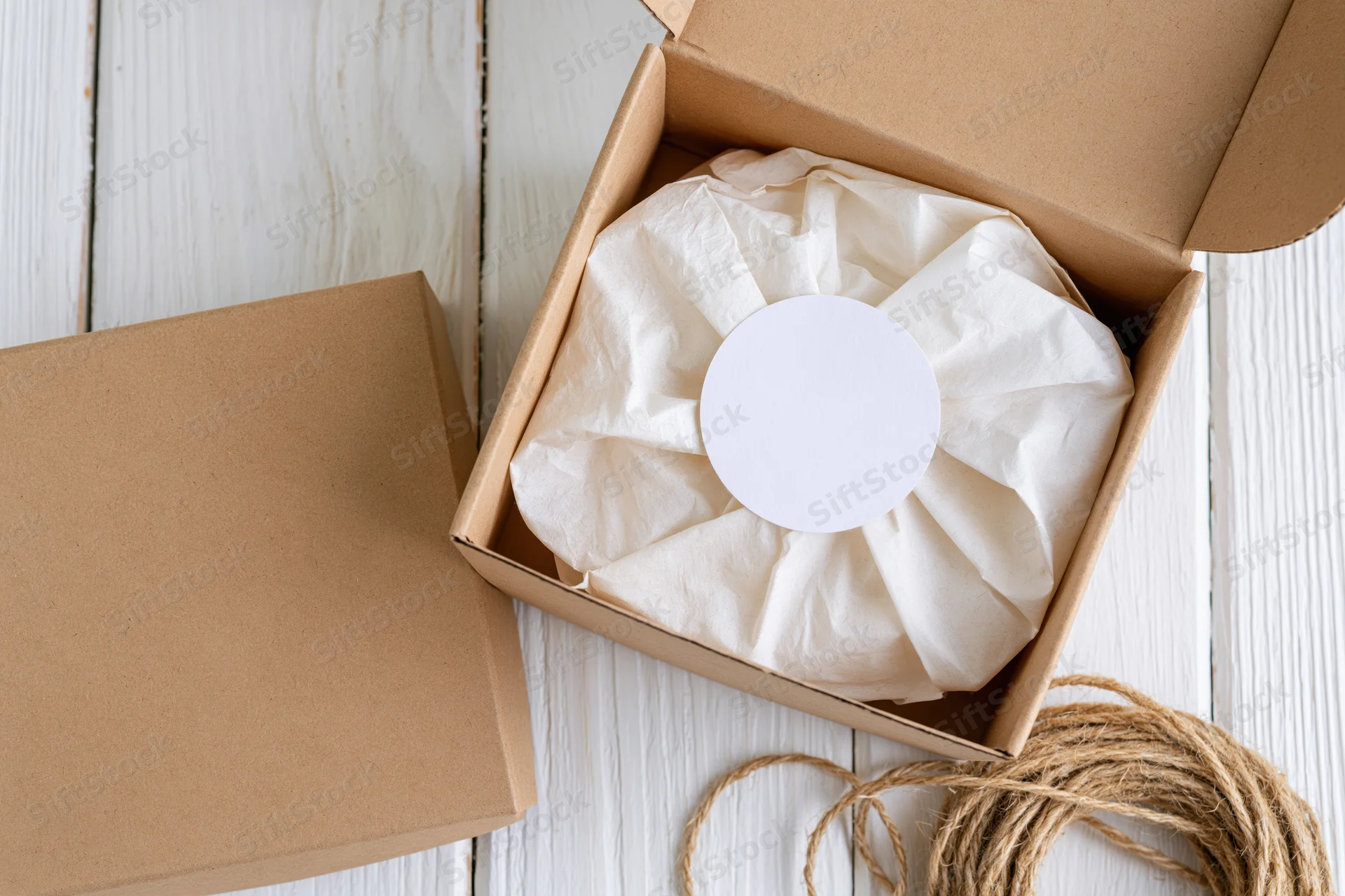 Open kraft cardboard gift box with white circular label on tissue paper, viewed from above on wooden surface with twine beside it