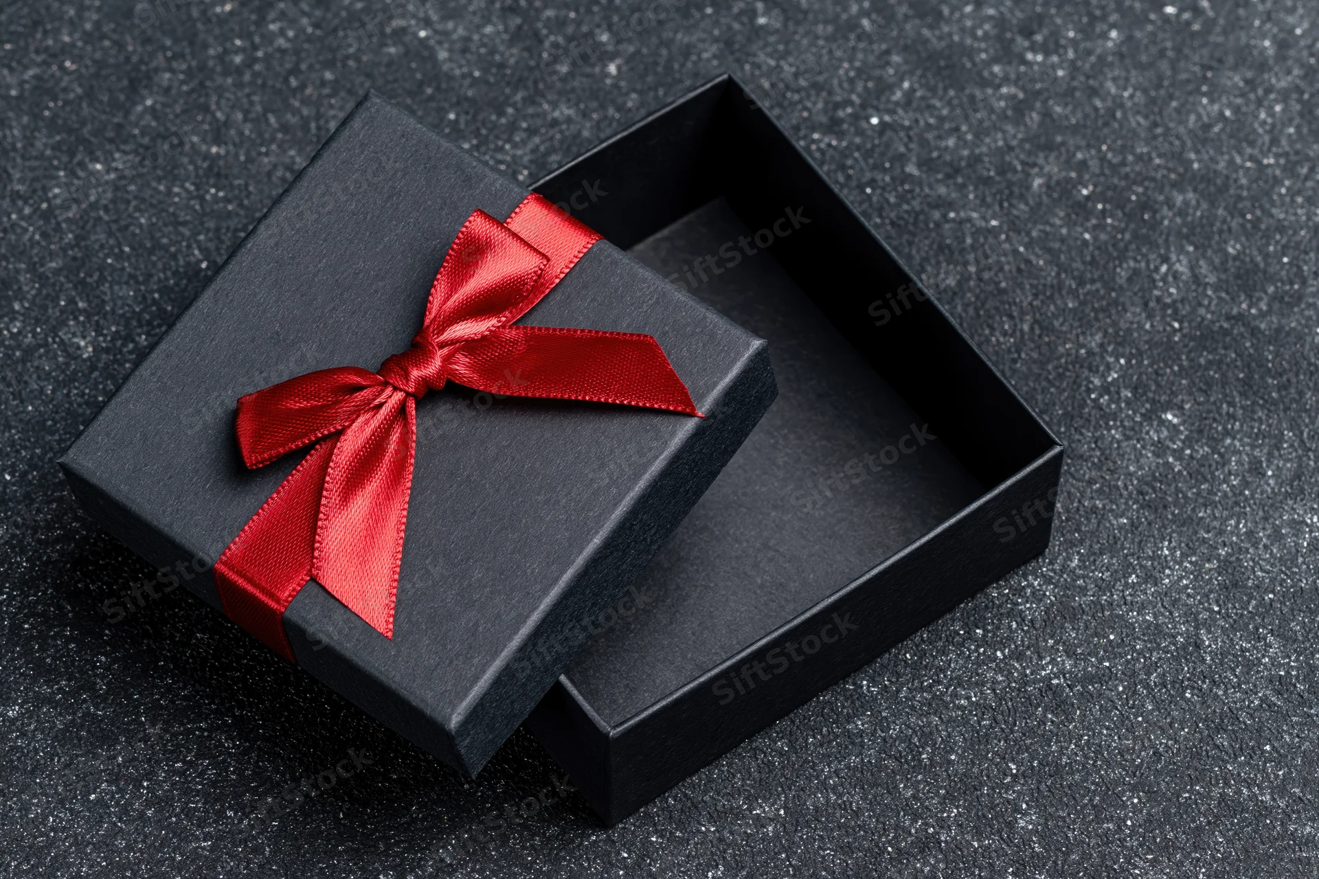 Open matte black gift box with red satin ribbon bow on dark textured surface