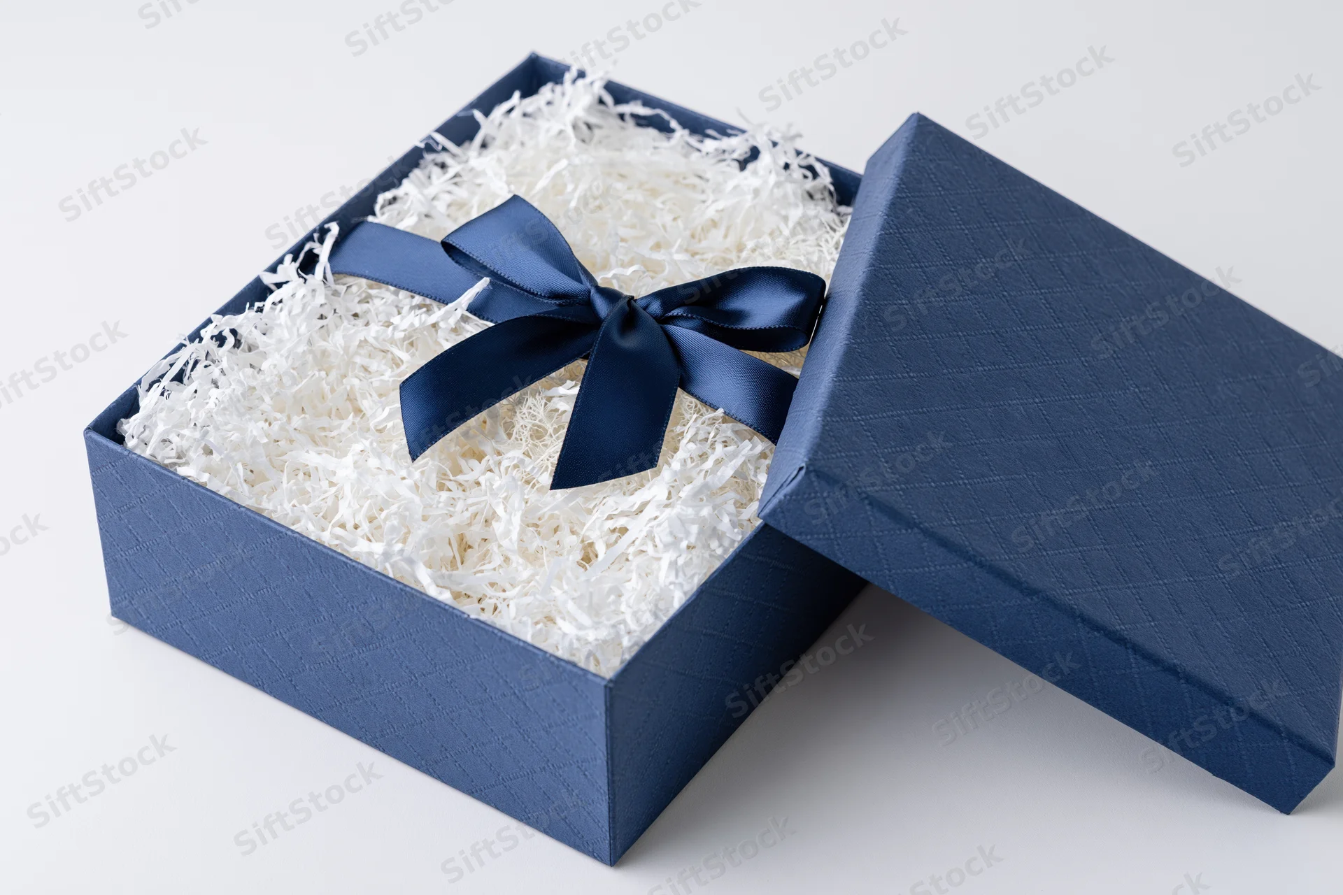 Open navy blue gift box with ribbon bow and shredded paper filling, lid leaning beside it on white surface