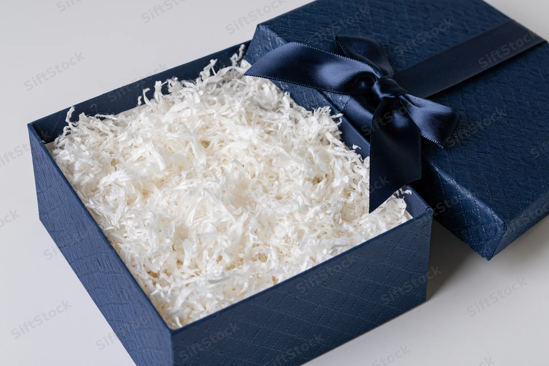 Open navy blue gift box with lid leaning against it filled with white shredded paper on white surface