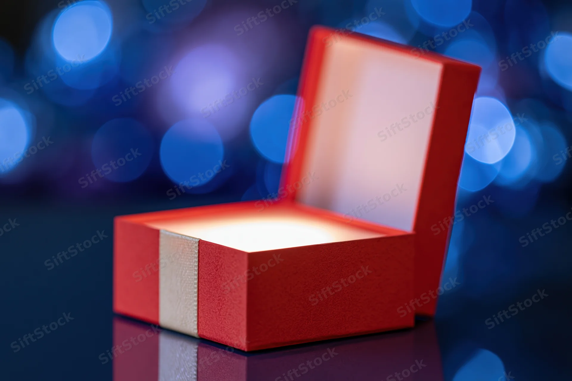 Open red gift box with glowing interior on reflective dark surface against blue bokeh background