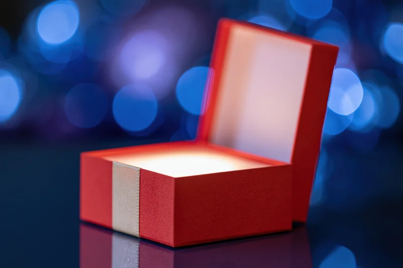 Open red gift box with glowing interior on reflective dark surface against blue bokeh background