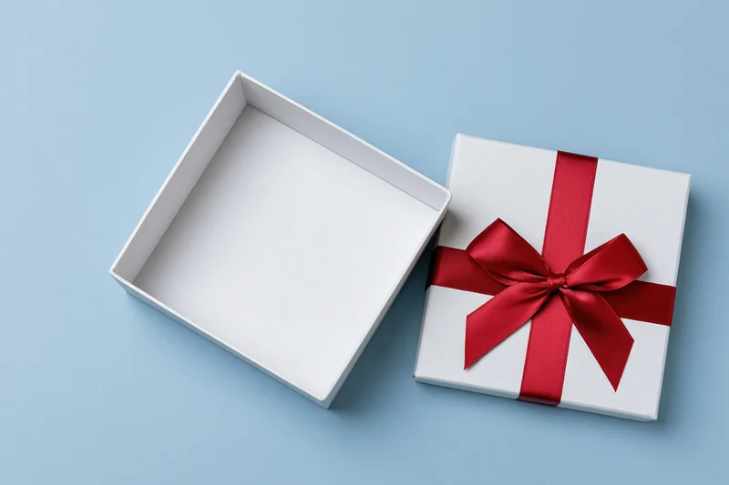 Open white gift box with red satin ribbon bow beside it on blue surface