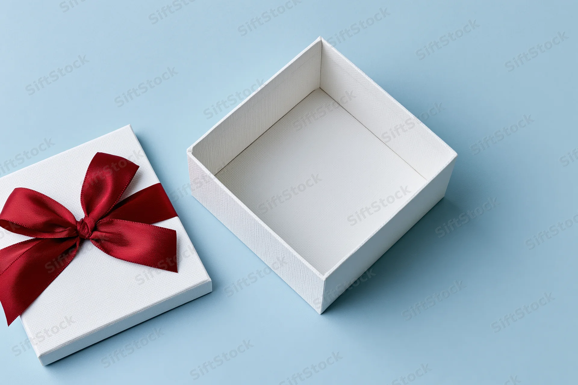 Open white gift box with red satin ribbon bow beside it on light blue surface