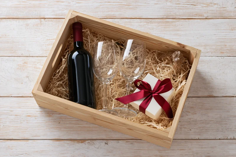 Wooden crate filled with wine bottle, two glasses and gift box tied with red ribbon on straw bedding over whitewashed wood surface
