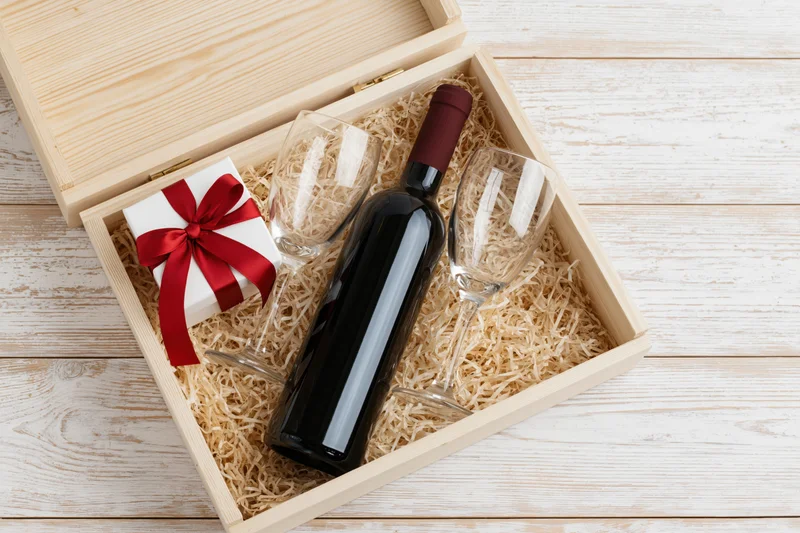 Open wooden gift box with wine bottle, two glasses and wrapped present nestled in straw on whitewashed wood surface
