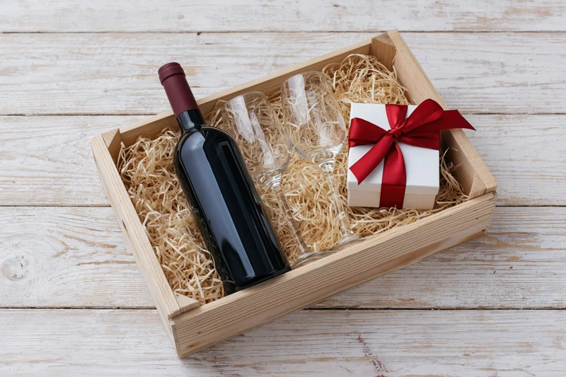 Wooden crate with wine bottle, two glasses and gift box wrapped in red ribbon, filled with straw on whitewashed wood surface