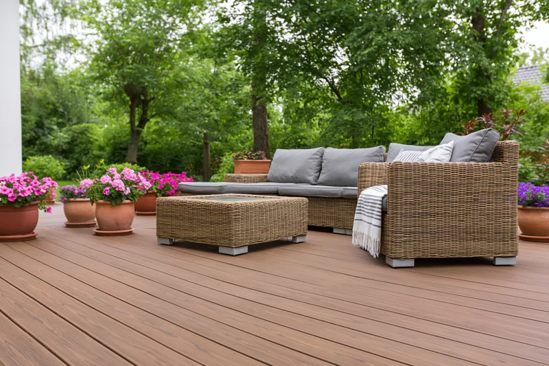Wicker sofa and armchair with coffee table on wooden deck surrounded by potted flowers and green trees