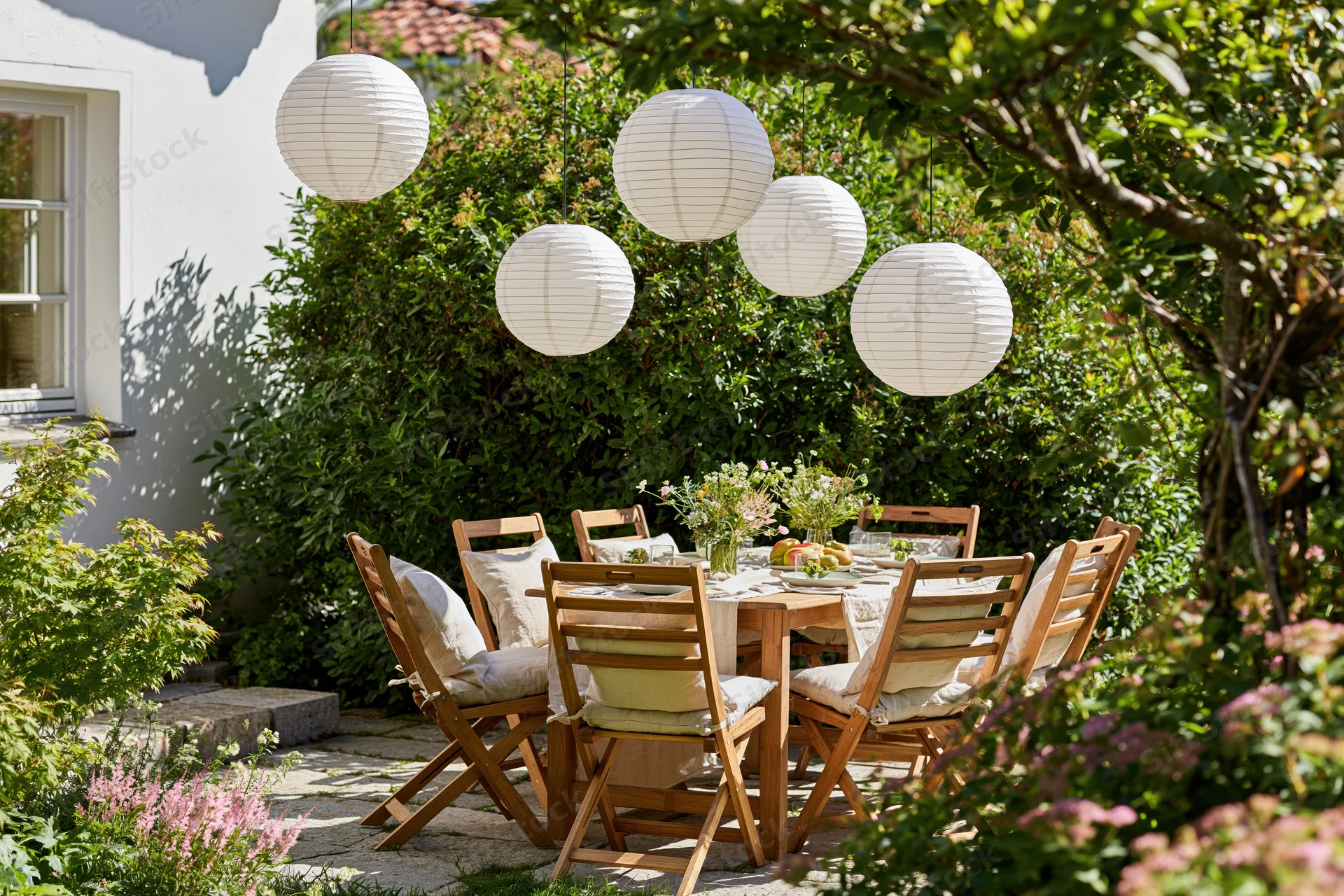 Wooden dining table set for a meal with hanging paper lanterns in a lush garden patio surrounded by greenery and flowers