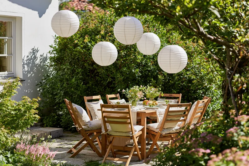 Wooden dining table set for a meal with hanging paper lanterns in a lush garden patio surrounded by greenery and flowers
