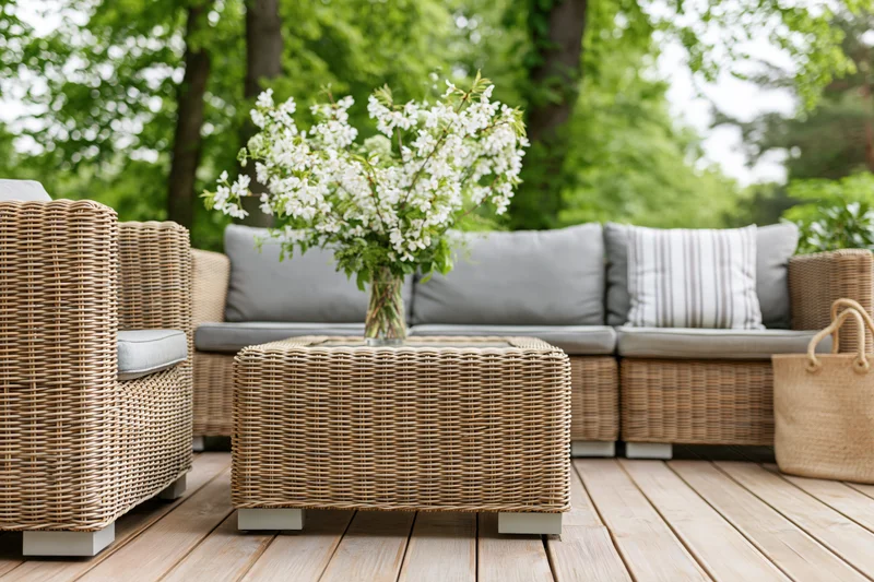 Wicker patio furniture set with cushions and flower vase on wooden deck surrounded by green trees
