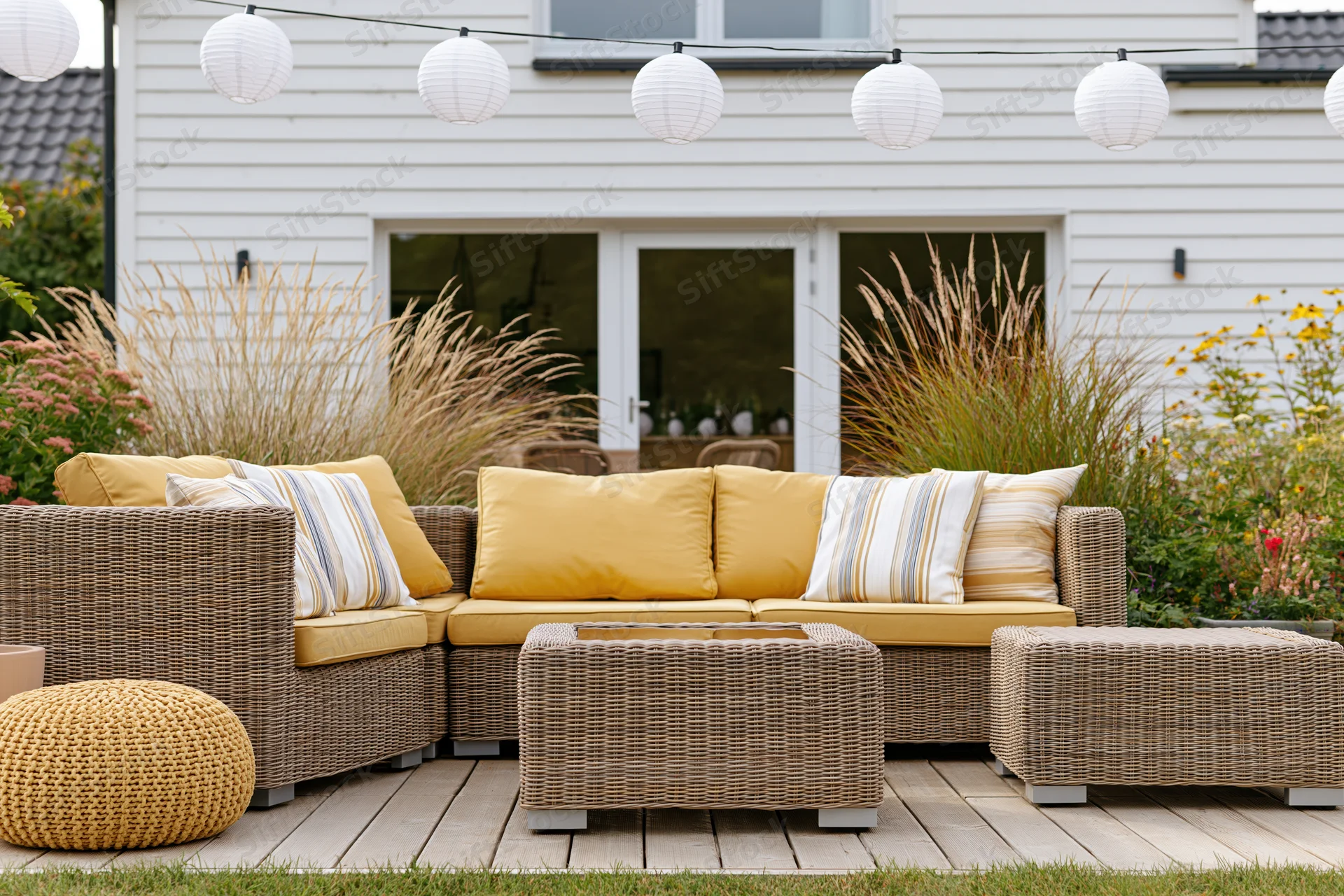 Wicker sectional sofa with yellow cushions and string lights on a wooden patio deck surrounded by ornamental grass and flowers