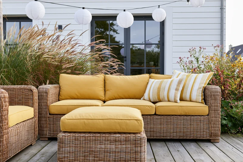 Wicker sofa and armchair with yellow cushions on a wooden deck surrounded by plants and string lanterns