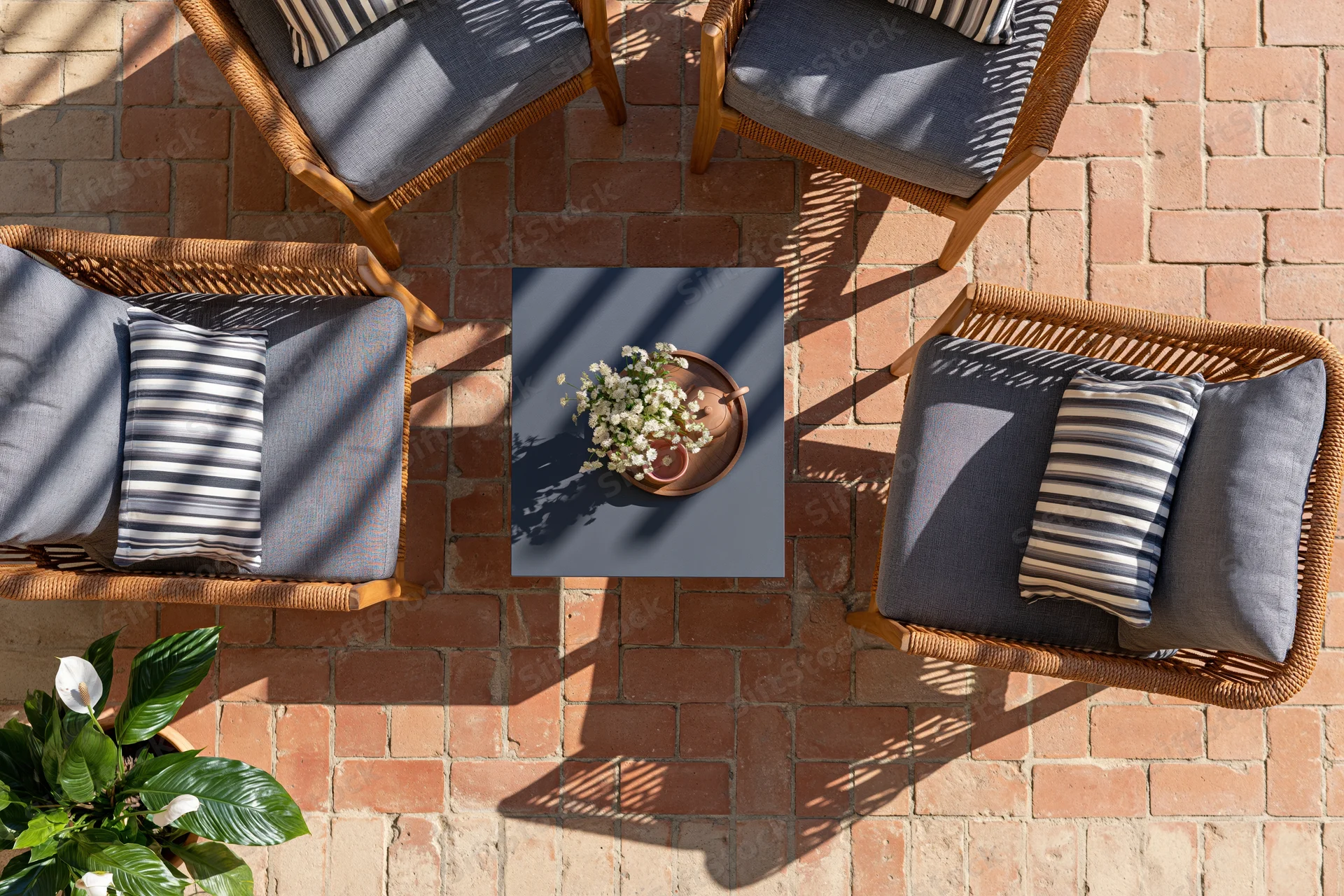 Overhead view of four wicker chairs with gray cushions and striped pillows arranged around a square table with white flowers on a brick patio floor