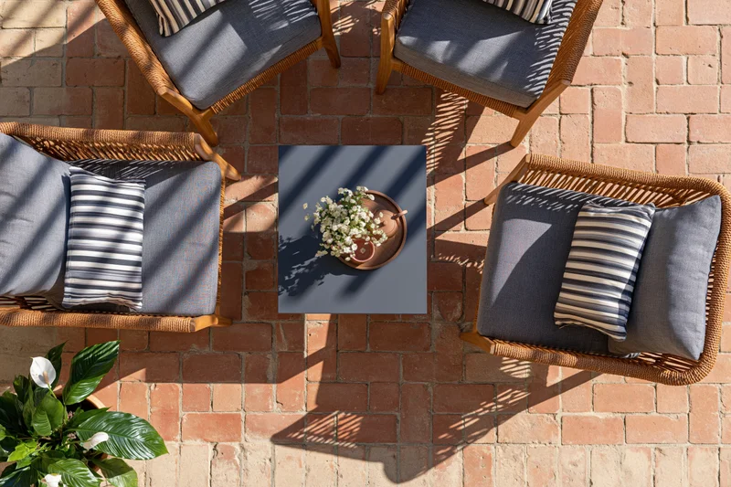Overhead view of four wicker chairs with gray cushions and striped pillows arranged around a square table with white flowers on a brick patio floor