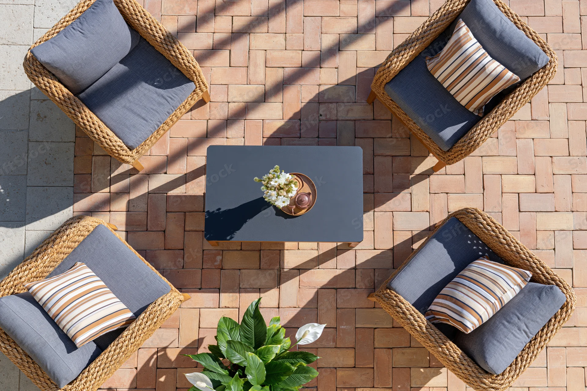 Overhead view of wicker patio chairs with gray cushions arranged around a coffee table on a brick patio with potted plant and sunlight shadows