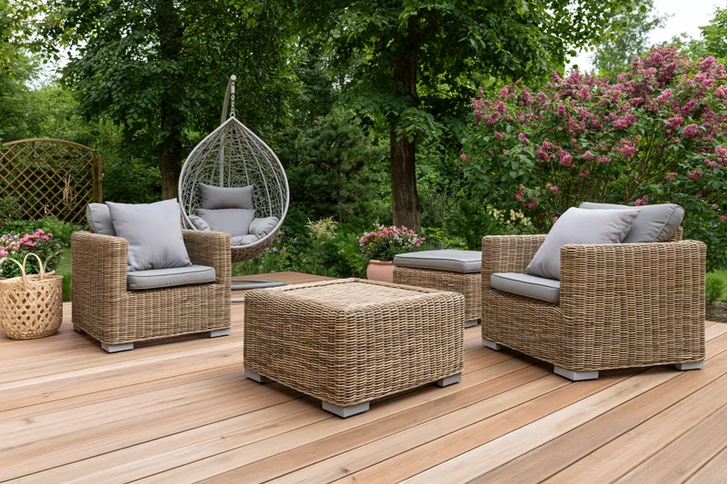 Wicker lounge chairs and ottoman arranged on a wooden deck surrounded by greenery and flowering shrubs