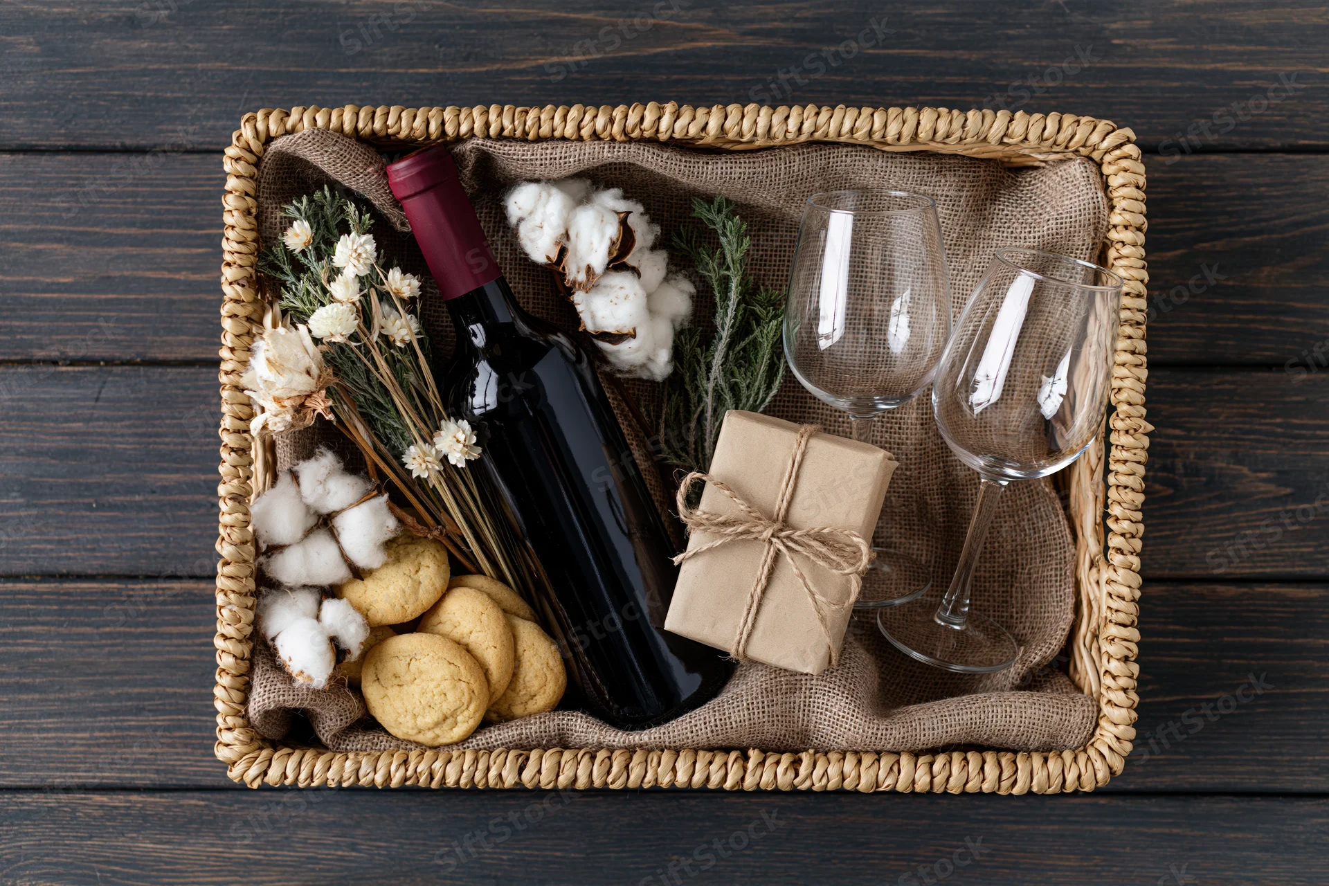 Woven wicker gift basket with wine, cookies, cotton, dried flowers and glasses on dark wood surface