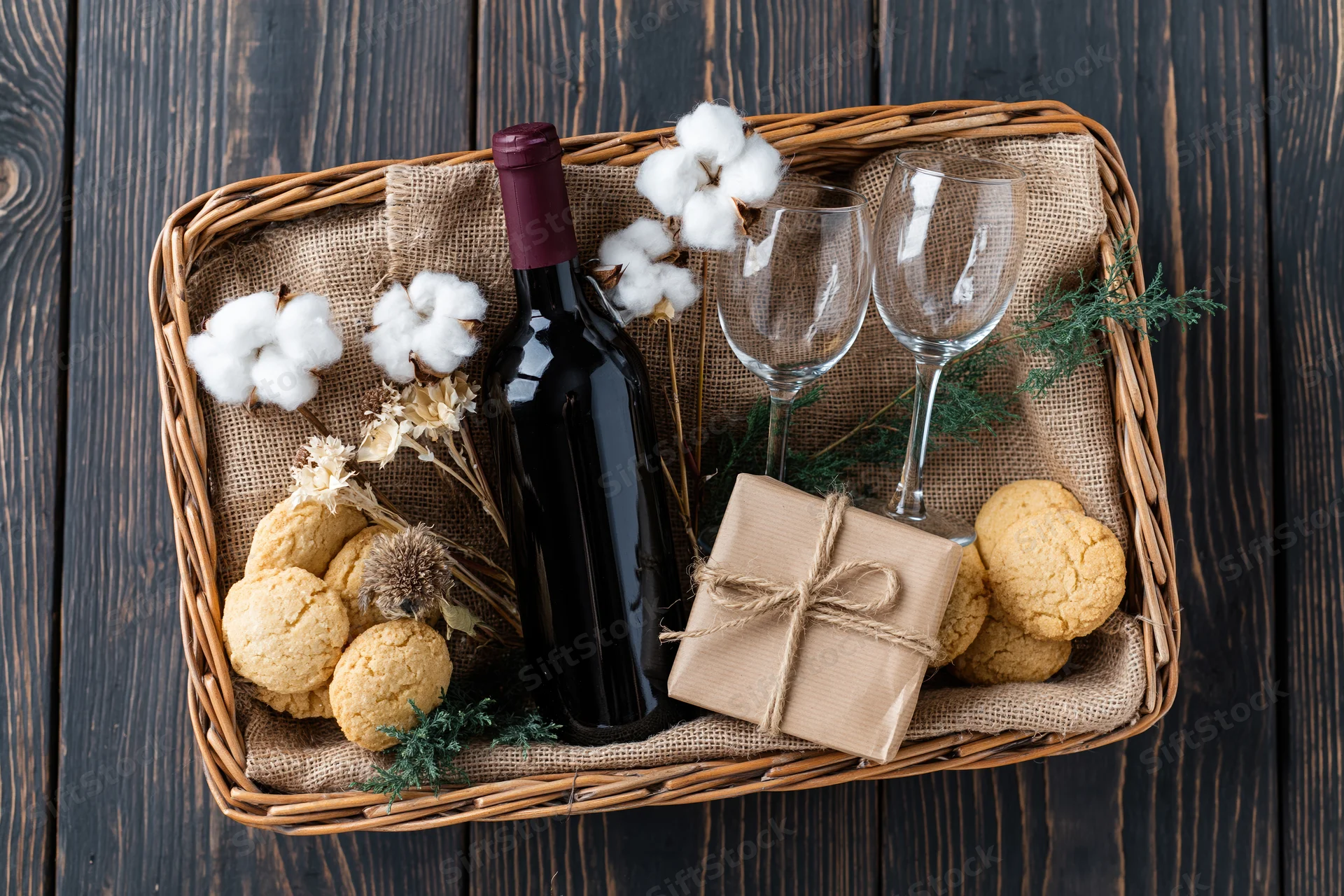 Overhead flat lay of a wicker gift basket with wine, cookies, cotton, and evergreen sprigs on dark wood