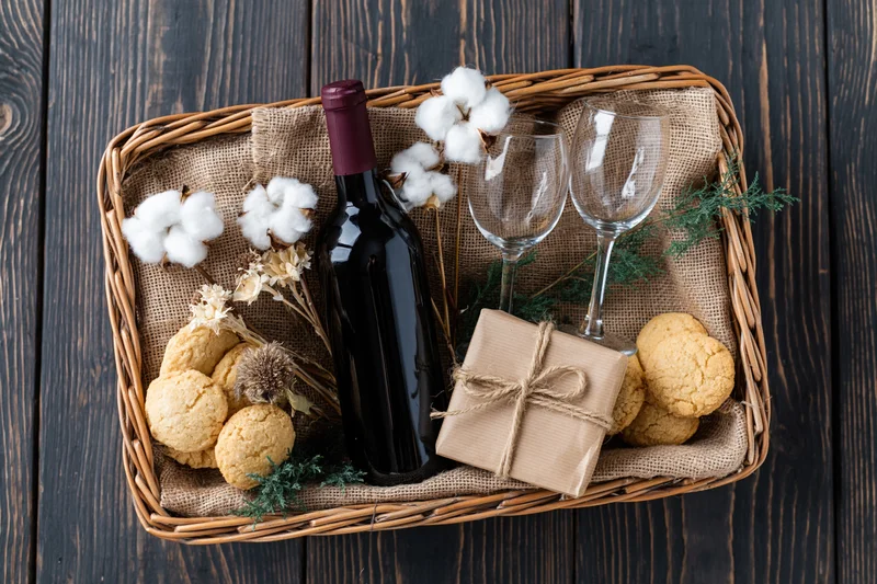 Overhead flat lay of a wicker gift basket with wine, cookies, cotton, and evergreen sprigs on dark wood