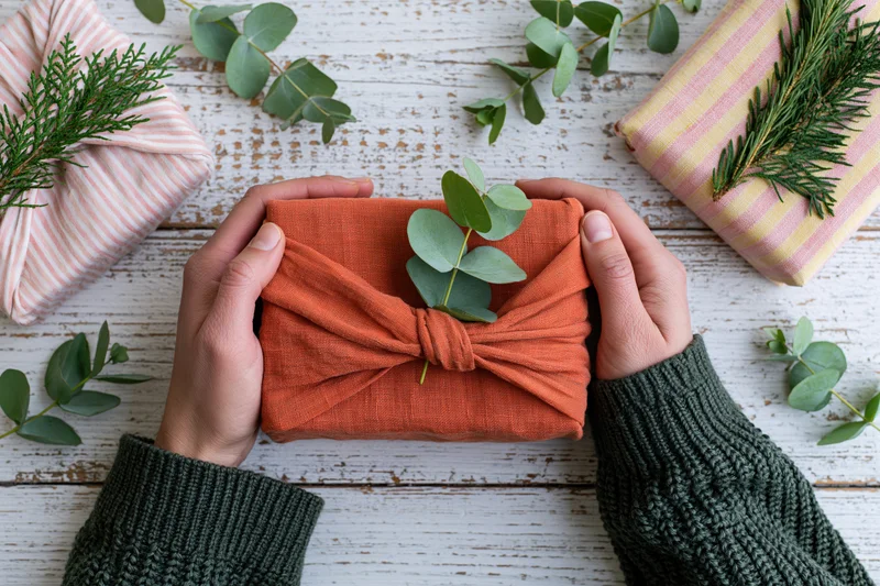 Hands holding a cloth-wrapped gift with eucalyptus sprig on a wooden table