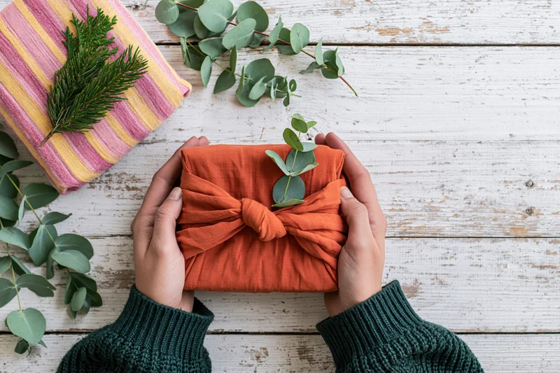 Hands holding a fabric-wrapped gift with eucalyptus sprig on a white wooden table