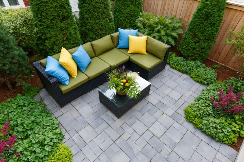 Overhead view of a wicker sectional sofa with colorful cushions on a stone patio surrounded by landscaped greenery and flowering plants