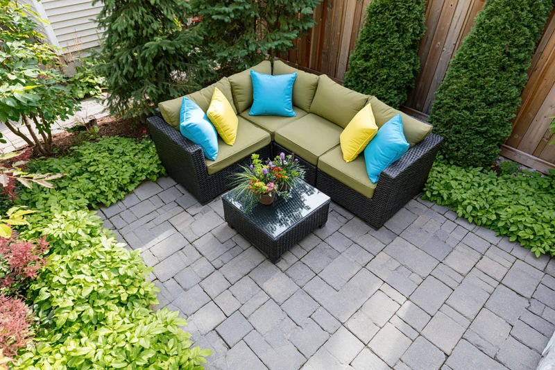 Overhead view of a wicker sectional sofa with colorful cushions on a paved patio surrounded by green shrubs and plants
