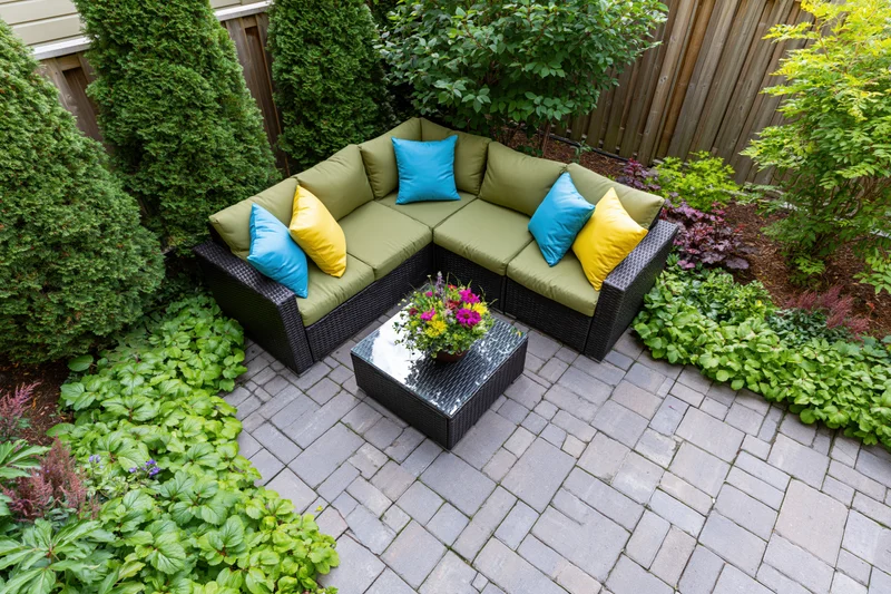 Overhead view of a dark wicker sectional sofa with colorful cushions on a brick patio surrounded by green shrubs and flowering plants in a small urban backyard
