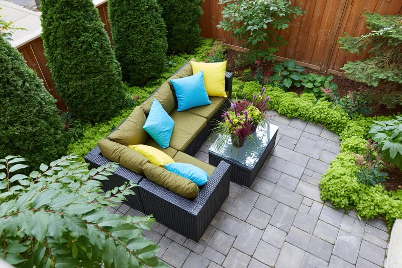 Overhead view of a small urban backyard patio with dark wicker sofa and colorful cushions surrounded by green plants and stone pavers