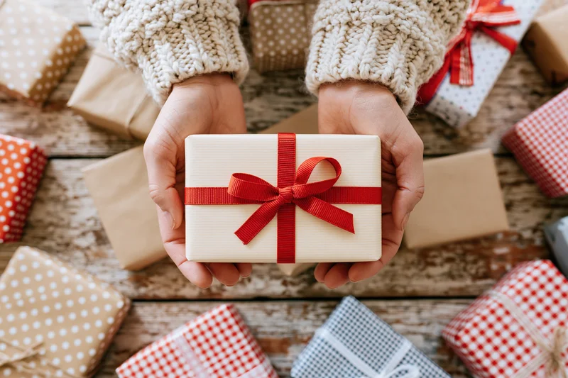 Hands holding a cream gift box with red ribbon over a wooden table surrounded by wrapped presents
