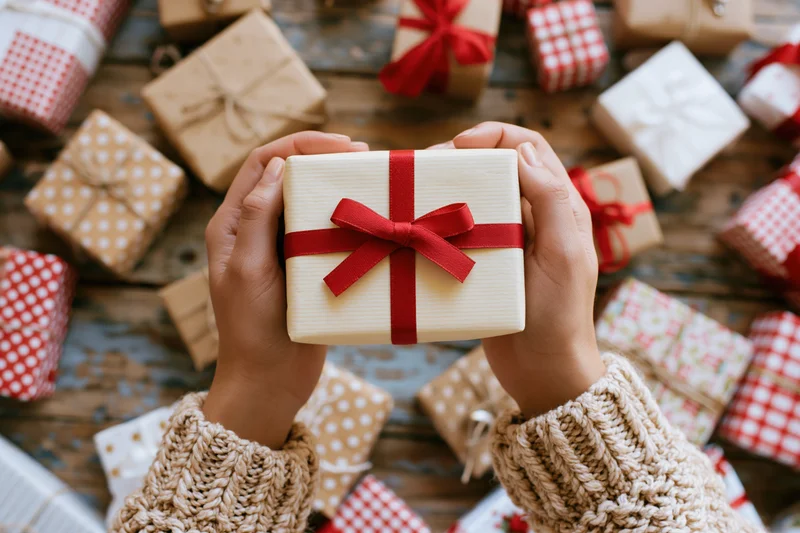 Hands holding a cream gift box with red ribbon over a wooden table surrounded by wrapped presents